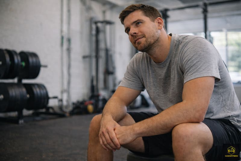 Sportlicher Mann sitzt nach dem Training entspannt auf einer Bank im Fitnessstudio als Symbol für Regeneration im Fitness-System und reflektierte Erholung nach Belastung